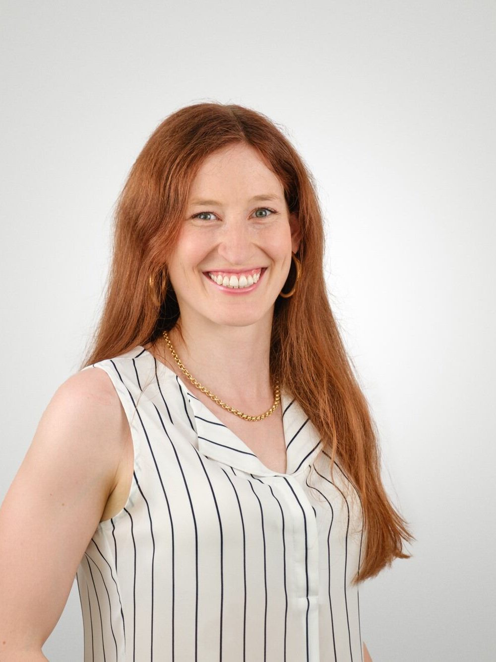 A smiling woman with long, wavy red hair wearing a sleeveless white blouse with black stripes and a gold chain necklace, posed against a light grey background.