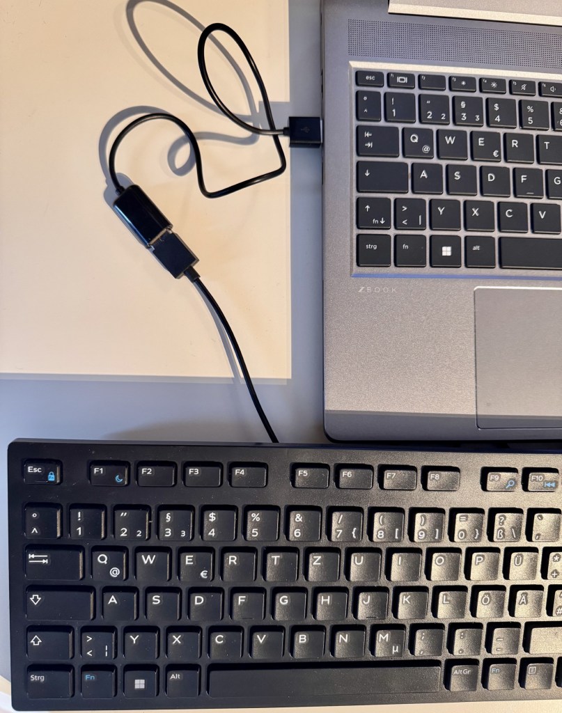 A top-down view of a black computer keyboard connected to a laptop via a USB cable.